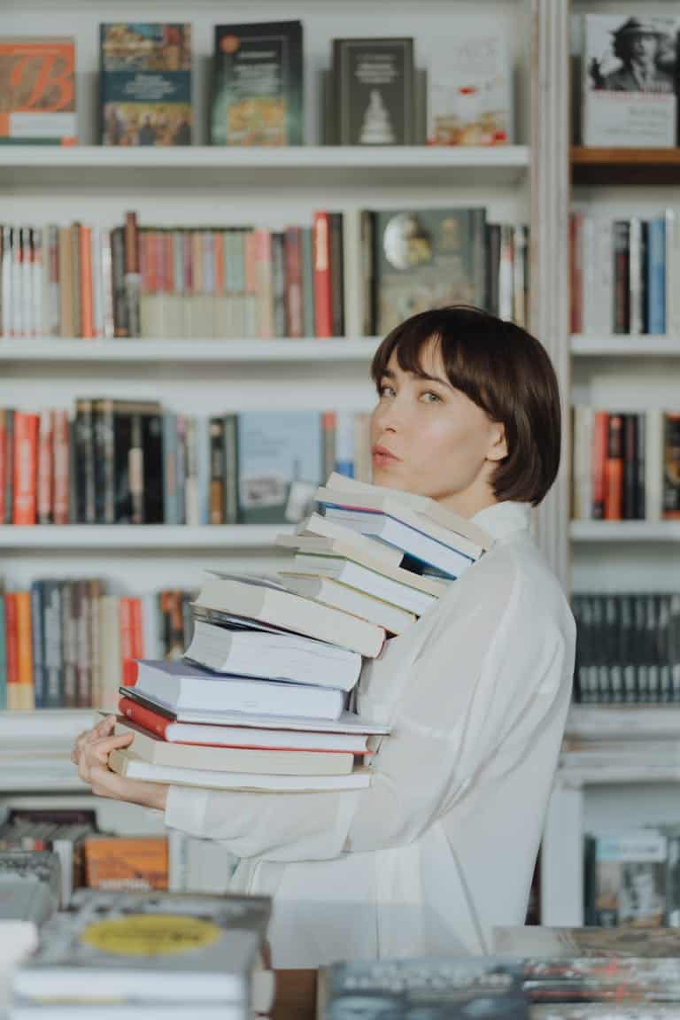 Woman in White Long Sleeve Shirt Carrying a Stack of Books