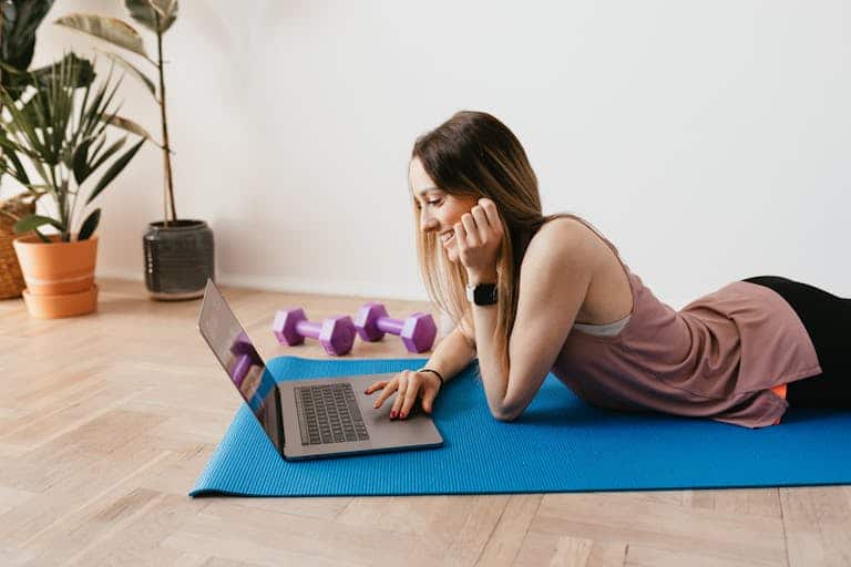 Side view of young happy slim female with long hair using laptop and smiling while lying on blue rubber mat