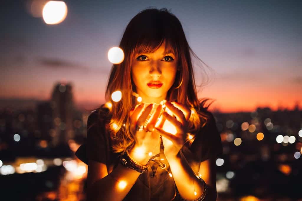 Romantic portrait of a woman holding glowing string lights against a city skyline at dusk.