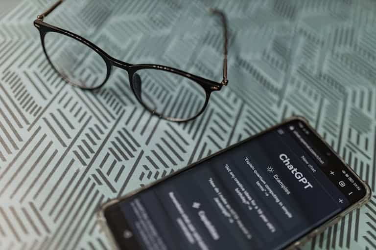 Eyeglasses next to a smartphone displaying the ChatGPT AI app on a patterned surface.